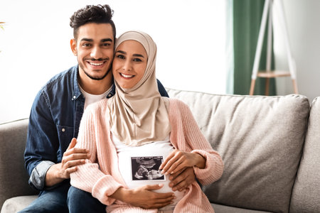 Happy Pregnant Muslim Family Sitting On Couch And Holding Baby Sonogram Pictureの写真素材