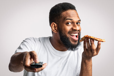Joyful African Millennial Man Eating Pizza Watching TV In Studioの写真素材