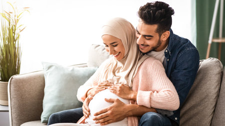 Happy islamic couple expecting baby hugging on couch at home