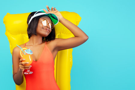Pretty black lady in swimsuit lying on inflatable lilo and enjoying tropical cocktail over blue studio backgroundの写真素材