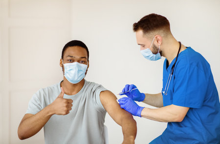 Black guy in face mask gesturing thumb up during covid-19 vaccination, approving of coronavirus immunization campaignの写真素材
