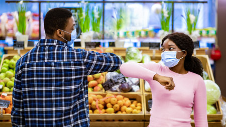 Millennial black woman and man in face masks greeting each other by touching elbows at supermarket during covid-19の写真素材
