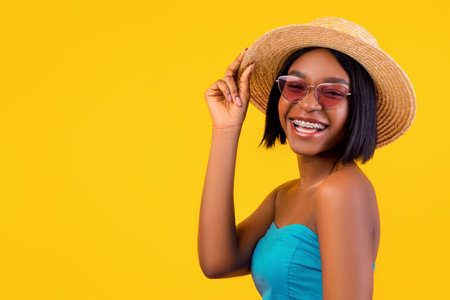 Smiling black lady in elegant dress and sunglasses touching her straw hat, ready for beach weekend...