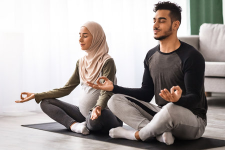 Expecting Muslim Spouses Practicing Yoga Together At Home, Sitting In Lotus Poseの写真素材