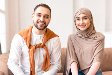 Muslim Couple Sitting On Sofa Smiling To Camera At Home