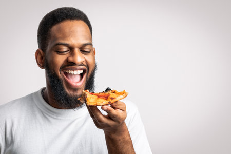 Joyful African Millennial Guy Eating Pizza Slice Over White Backgroundの写真素材
