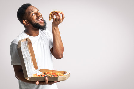 Hungry African Guy Eating Slice Of Pizza Posing In Studioの写真素材