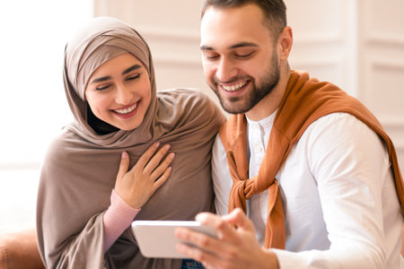 Muslim Couple Using Smartphone And Laughing Sitting In Living Roomの写真素材