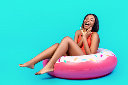 Portrait of excited black lady in bikini touching her face, sitting in donut shaped inflatable ring...