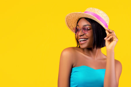 Portrait of cheerful black lady with dental braces wearing summer dress, sunglasses and straw hat on...