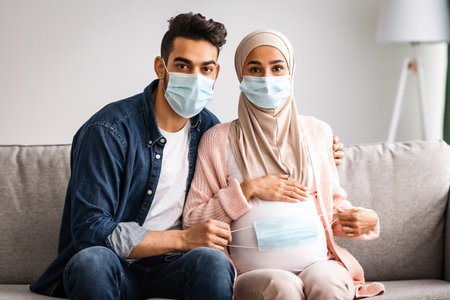 Pregnant Muslim Couple Sitting On Couch Holding Protective Medical Mask Near Bellyの写真素材