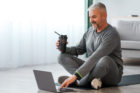 Senior man in sportswear sitting on fitness mat, using laptopの写真素材
