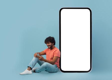 Rlaxed young indian man using cell phone, sitting on blueの写真素材