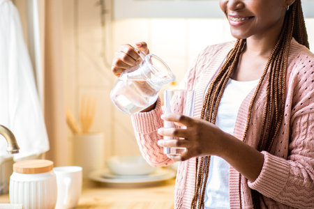 Young African American Lady Drinking Water In Kitchen, Holding Jug And Glassの写真素材