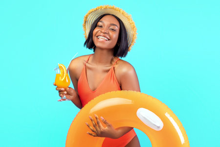 Happy African American lady with inflatable ring and tasty tropical cocktail smiling at camera on blue studio backgroundの写真素材
