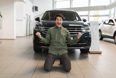 Excited young guy shaking fists, making YES gesture, standing on his knees after buying new auto at dealershipの写真素材