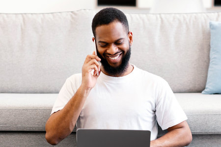 Black Freelancer Guy Talking On Phone Using Laptop At Home
