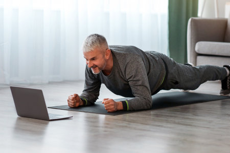 Athletic mature bearded man doing exercises at home, using laptopの写真素材