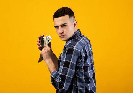 Young man with wallet full of money being greedy over his wealth on orange studio backgroundの写真素材