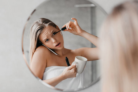 Busy young lady talking on smartphone, applying mascara and drinking coffee simultaneously in a hurryの写真素材