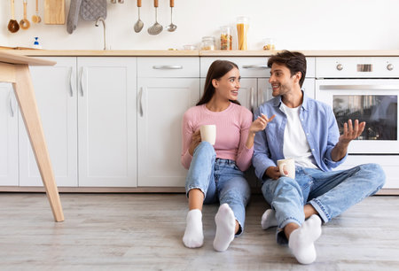Smiling millennial european woman and man with cups of drink, sitting on floor, talk in kitchen interiorの写真素材