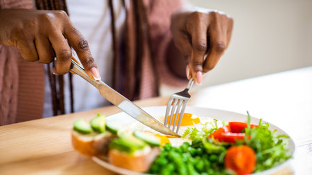 Unrecognizable african woman eating tasty breakfast in kitchen, using fork and knifeの写真素材