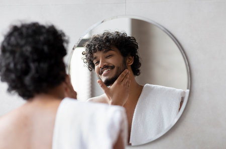 Handsome indian man looking in mirror in bathroom and touching beard, enjoying morning beauty routineの写真素材