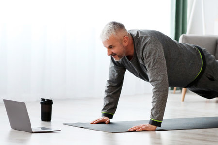 Sporty mature bearded man doing exercises at home, using laptopの写真素材