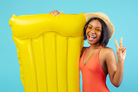 Cheerful black lady in swimsuit holding yellow lilo and showing peace gesture on blue studio...