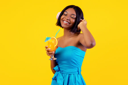 Black lady in casual outfit holding tropical summer cocktail and listening to music in headphones on orange backgroundの写真素材