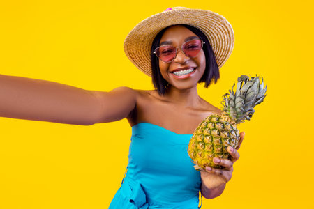 Sexy black lady in trendy summer dress taking selfie with pineapple on orange studio background