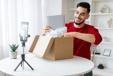 Handsome Arab Male Blogger Unboxing Parcel With Laptop On Cameraの写真素材