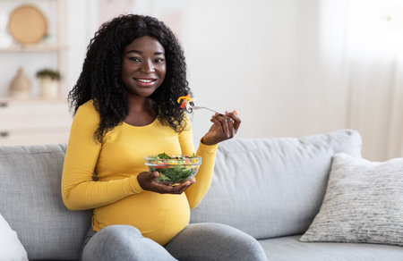 Smiling pregnant black woman eating fresh salad, sitting on sofaの写真素材
