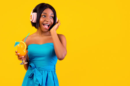 Summer clubbing. Black lady with tropical cocktail wearing headphones, listening to dance music on orange backgroundの写真素材