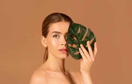 Portrait of pretty young woman with smooth skin and nude makeup closing her eye with monstera leaf on brown backgroundの写真素材