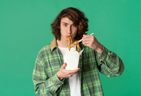Food delivery concept. Young man eating takeaway noodles on color background, blank space for designの写真素材