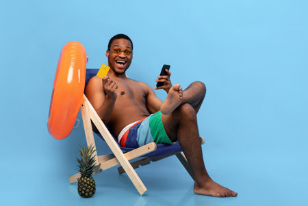 Excited black man holding credit card and smartphone, shopping online in lounge chair on blue studio backgroundの写真素材