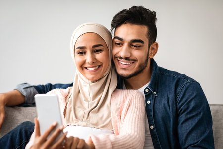 Cheerful Pregnant Middle Eastern Spouses Browsing Internet On Smartphone Together At Homeの写真素材