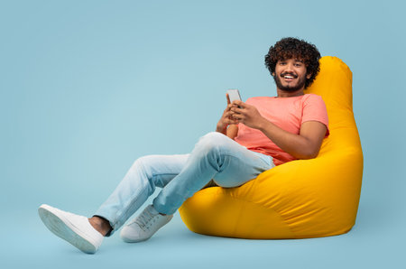 Relaxed eastern guy chilling at bean bag with smartphoneの写真素材