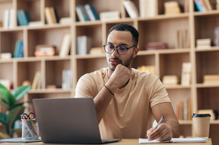 Focused arab man in glasses using laptop sitting at desk and writing in notebook, watching webinar studying onlineの写真素材