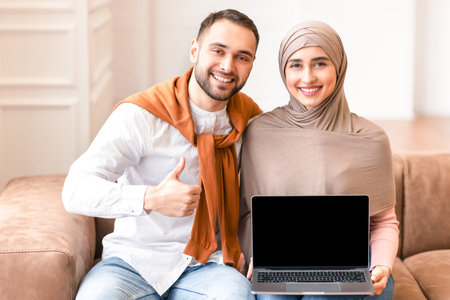 Muslim Couple Showing Laptop Screen Sitting On Couch At Homeの写真素材