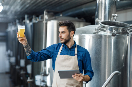 Young guy worker in apron with tablet checks color of lager in glass in interior