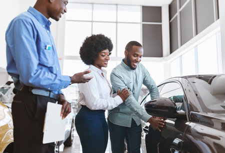 Car sales business. Manager talking to Afro couple, showing them new auto at dealership shopの写真素材