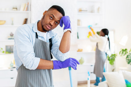 Young black guy leaning on mop, feeling bored or tired of cleaning home with his girlfriend, indoorsの写真素材