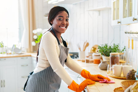 Young black lady in apron and rubber gloves wiping kitchen table, her boyfriend washing dishes on...