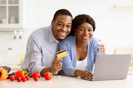 Happy black couple using laptop and credit card in kitchenの写真素材