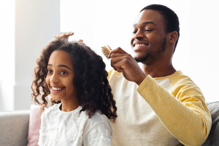 Black father combing daughter hair while sitting behind on couchの写真素材