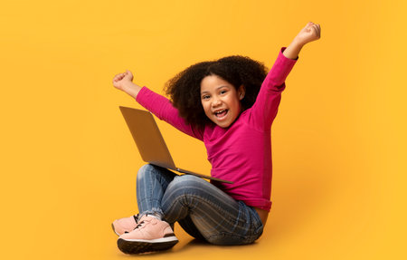 Joyful little black girl sitting on floor celebrating success with laptopの写真素材