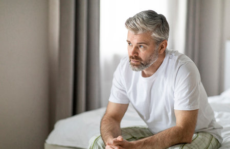 Pensive mature caucasian man sitting on bed at homeの写真素材