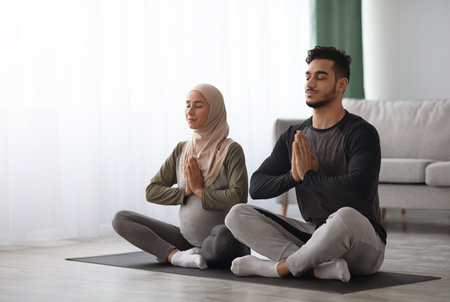 Pregnant Muslim Woman In Hijab Meditating Together With Her Husband At Homeの写真素材
